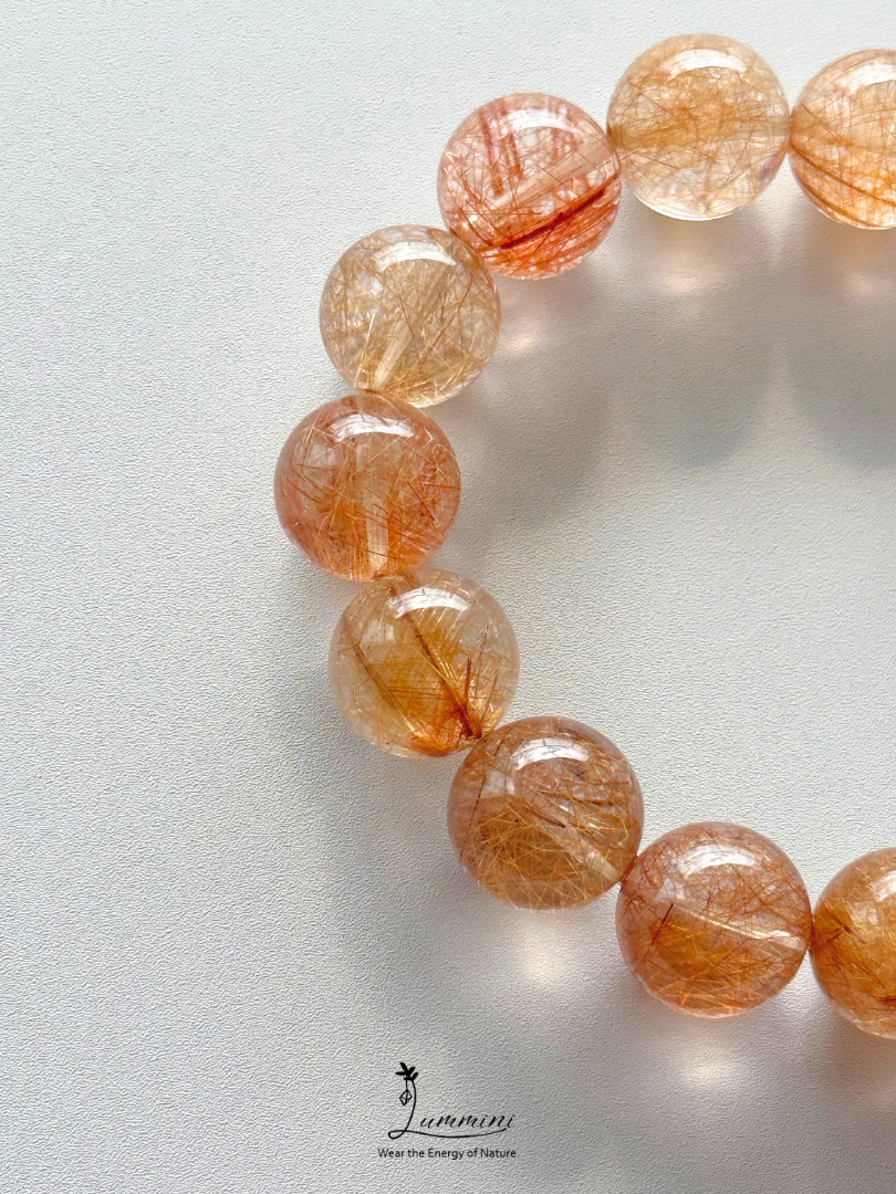 A close-up of a beaded bracelet made of translucent round beads filled with fine golden-orange hair-like inclusions, photographed on a light neutral background. The bracelet is cropped at the top and right edges, with a small 'Jummin' logo visible near the bottom center.
