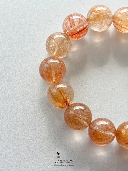 A close-up of a beaded bracelet made of translucent round beads filled with fine golden-orange hair-like inclusions, photographed on a light neutral background. The bracelet is cropped at the top and right edges, with a small 'Jummin' logo visible near the bottom center.