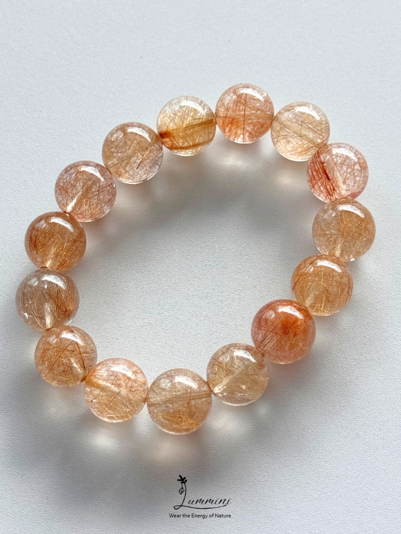 A beaded stretch bracelet made of round, translucent peach and orange gemstone beads with fine metallic-looking inclusions, photographed on a light gray background. A small 'Jummin' logo watermark is visible at the bottom.
