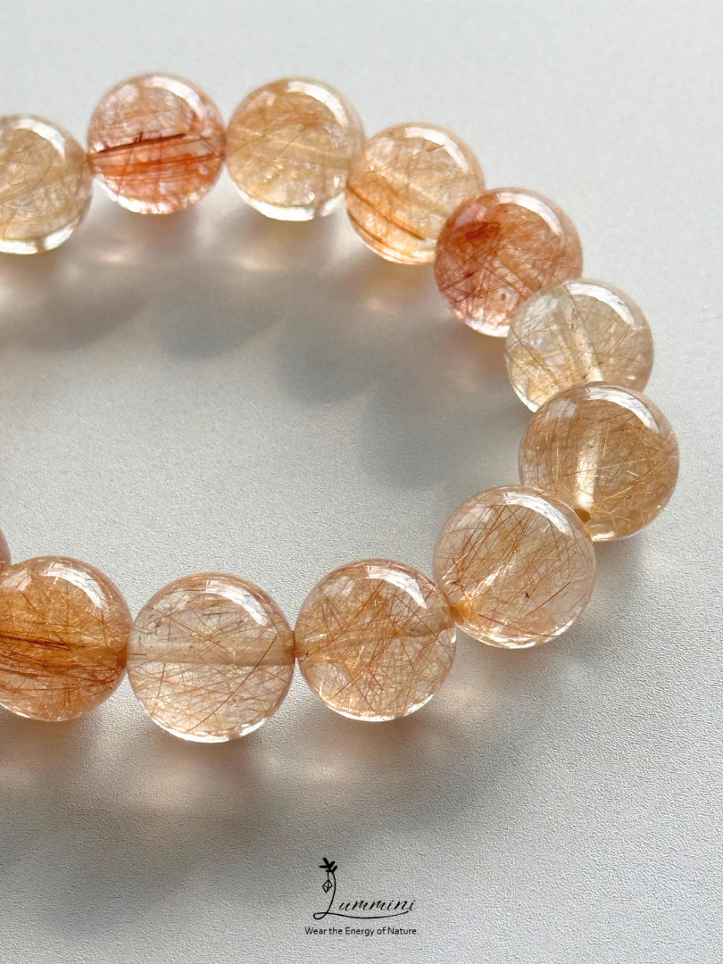 A beaded bracelet made of translucent round beads filled with fine golden-white hair-like inclusions, photographed on a light gray background. A small 'Jumino' logo appears near the bottom center.