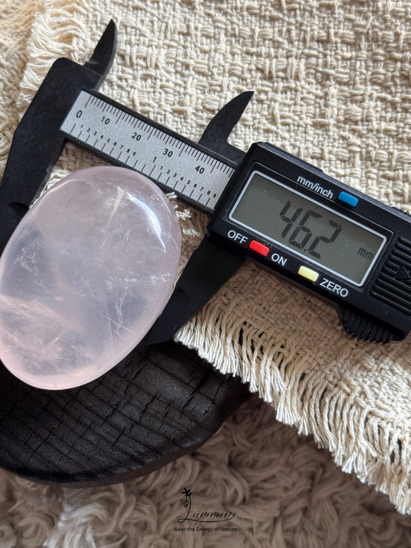 A rose quartz crystal with a smooth, polished oval shape is shown under a digital caliper displaying 46.2 mm, resting on a black textured surface over a beige fringed fabric.